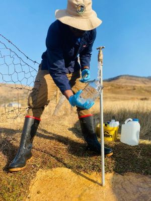 Person filling up jar with water from a water tap coming out of the ground in a rural setting
