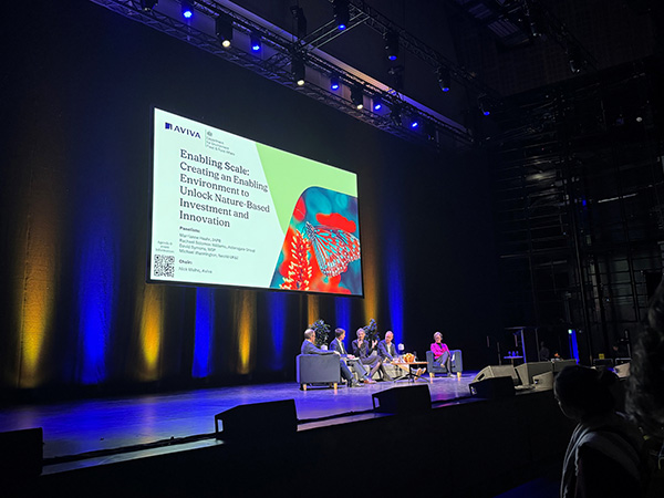 Photograph of the panel discussion at the Aviva/Defra Business & Finance Day. A group of 5 of 6 panel members are seated in a semi-circle. Behind and above them is a large screen displaying a presentation