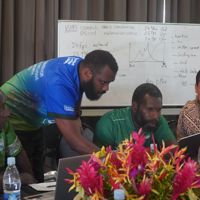 4 people sat along a table with another person leaning over showing them something on a laptop